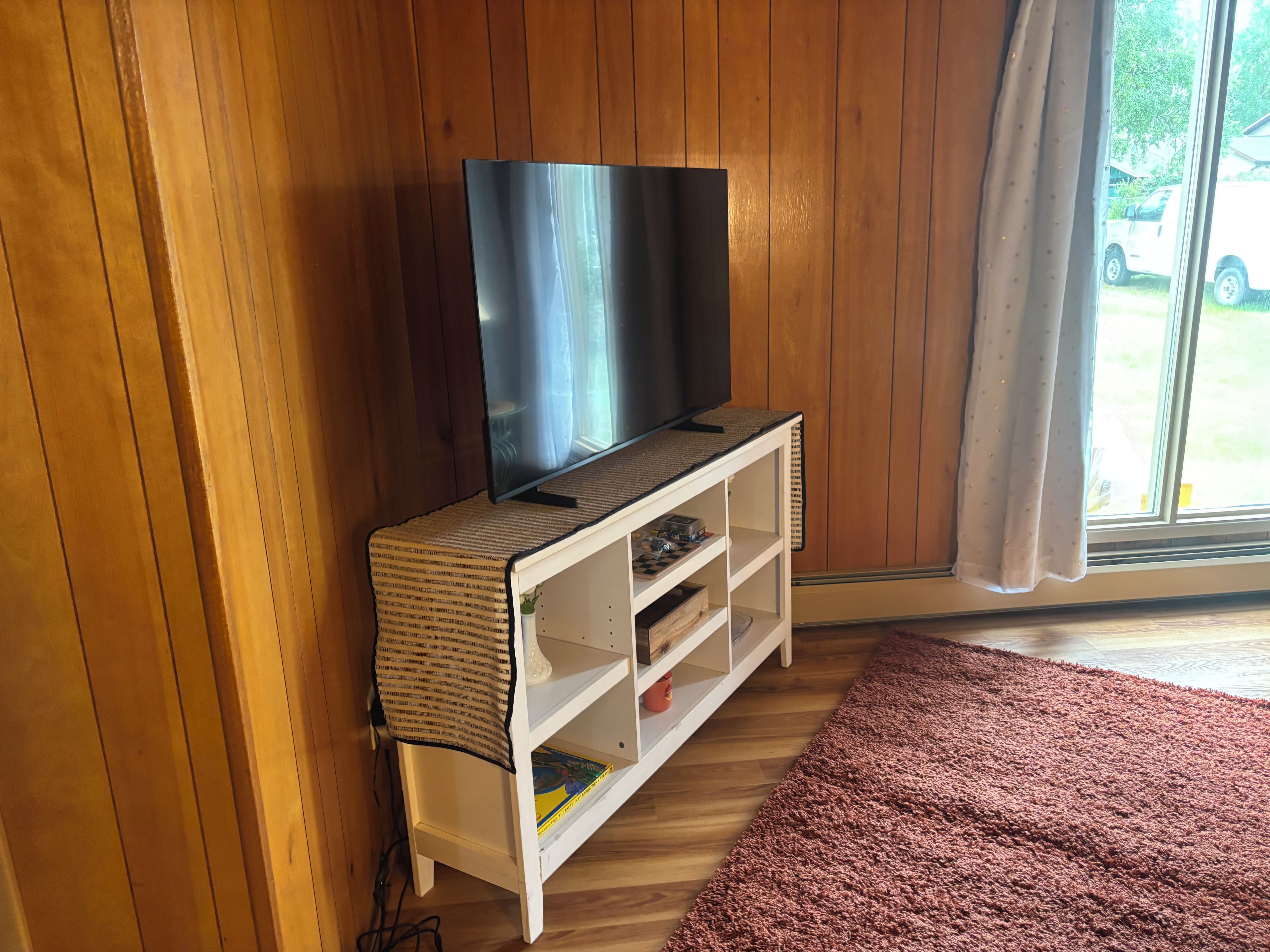 A room with wooden walls, a flat-screen TV on a white TV stand, a window with a view of a parked car, and a brown carpet.