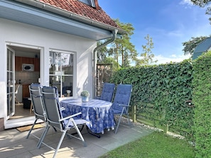 Eine Terrasse mit Tisch und Stühlen, eine durch eine offene Tür erkennbare Küche und ein grüner Heckenstreifen.