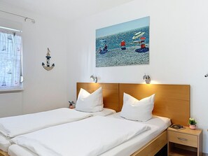 Ein Doppelbett mit weißer Bettwäsche, ein Holzkopfteil, ein kleiner Nachttisch mit Lampe und ein gerahmtes Bild mit Strandmotiv.