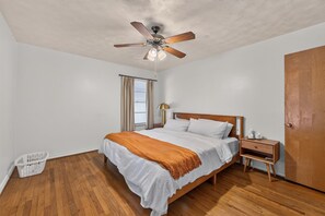 Ein Schlafzimmer mit einem hölzernen Bett, einem Nachttisch, einer Lampe, einem Deckenventilator und einem Fenster mit Vorhängen.