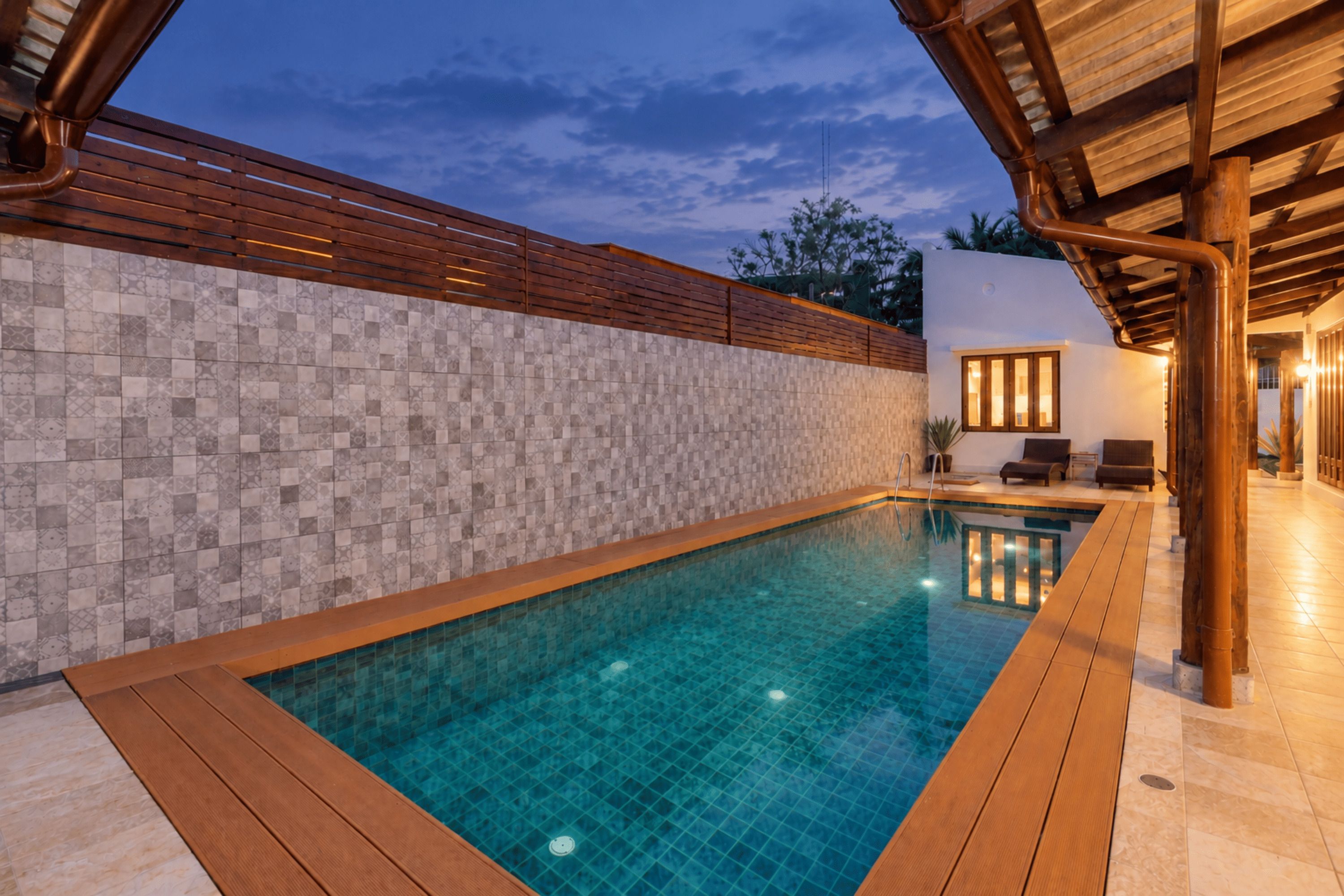 Une piscine avec une terrasse carrelée, une clôture en bois et un espace détente couvert offrant une vue sur le ciel.