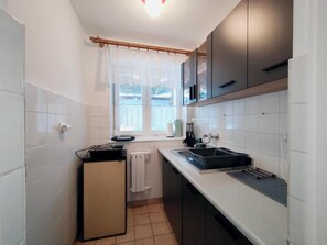 A compact kitchen with white tiled walls, a small refrigerator, a sink, and a window with a curtain.