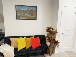 A living room with a black sofa, colorful cushions, a framed painting, and a potted plant. - Private Suite Near Calgary Airport (Calgary)