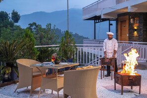 An outdoor dining area with wicker furniture, a table set with plates and glasses, and a chef cooking on a grill. - ELIVAAS Signature SECRET ESCAPE SWARAN (Solan)