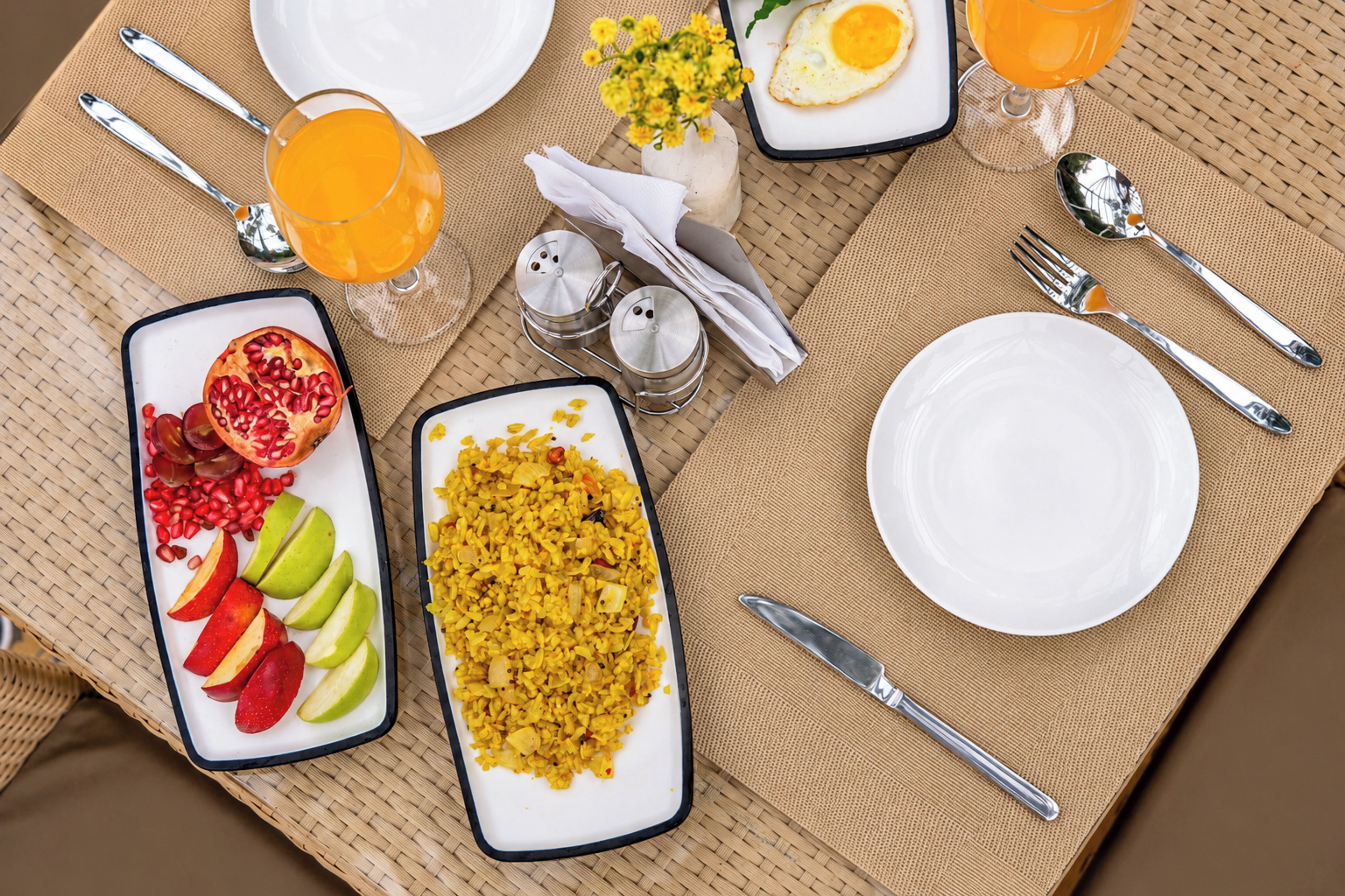 Un desayuno con un plato de fruta, un tazón de arroz amarillo, un vaso de jugo de naranja y una taza de café.