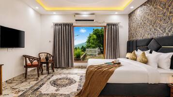 A hotel room with a bed, a TV, a desk with a chair, a window with curtains, and a view of greenery.