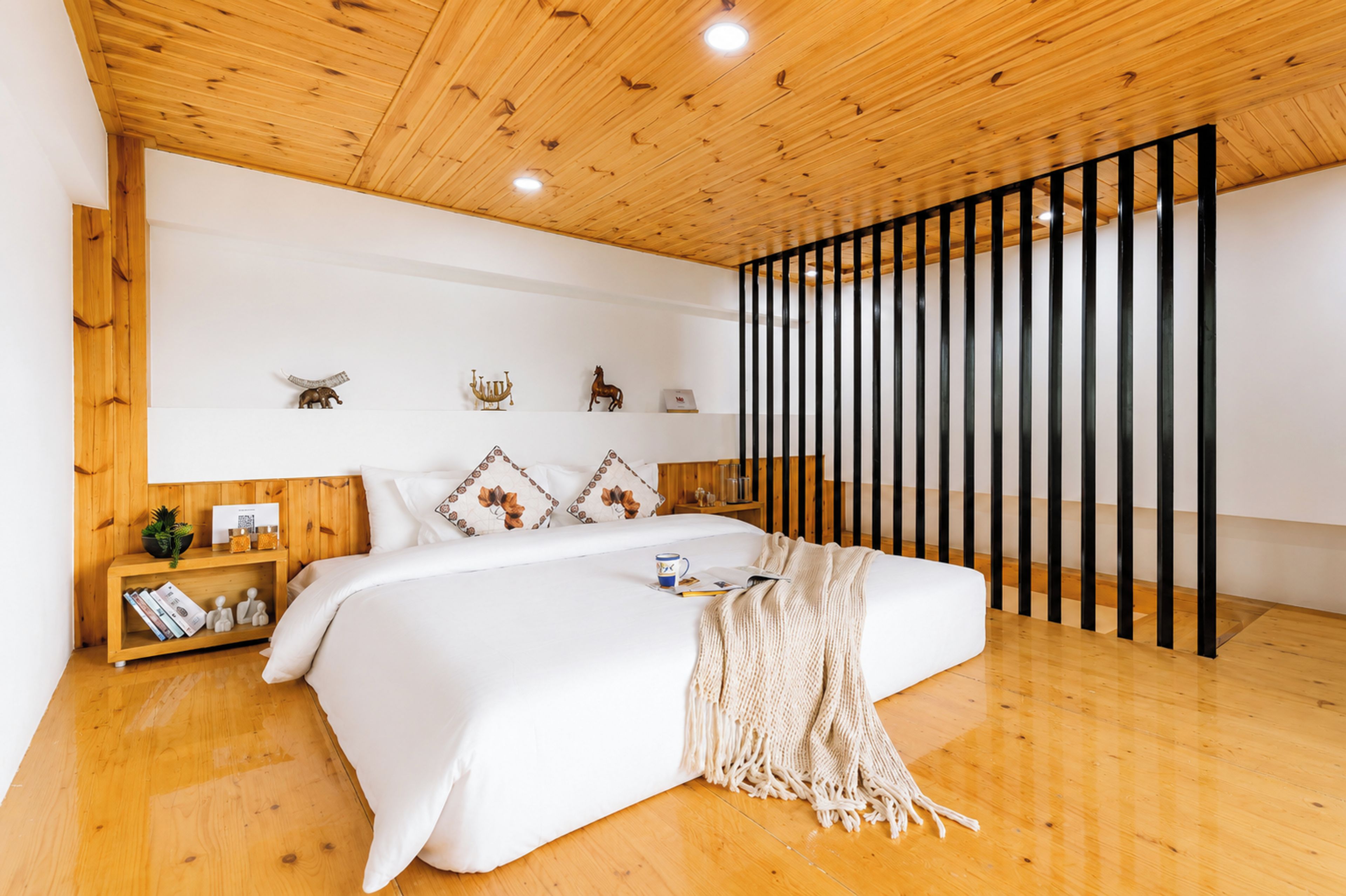 Une chambre avec un plafond en bois, un lit recouvert de linge de lit blanc, une table de chevet avec des livres et une plante, et une étagère murale ornée d’objets décoratifs.
