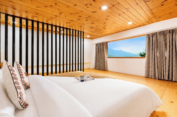A bedroom with a large bed, wooden ceiling, black railing, and a window with a mountain view.