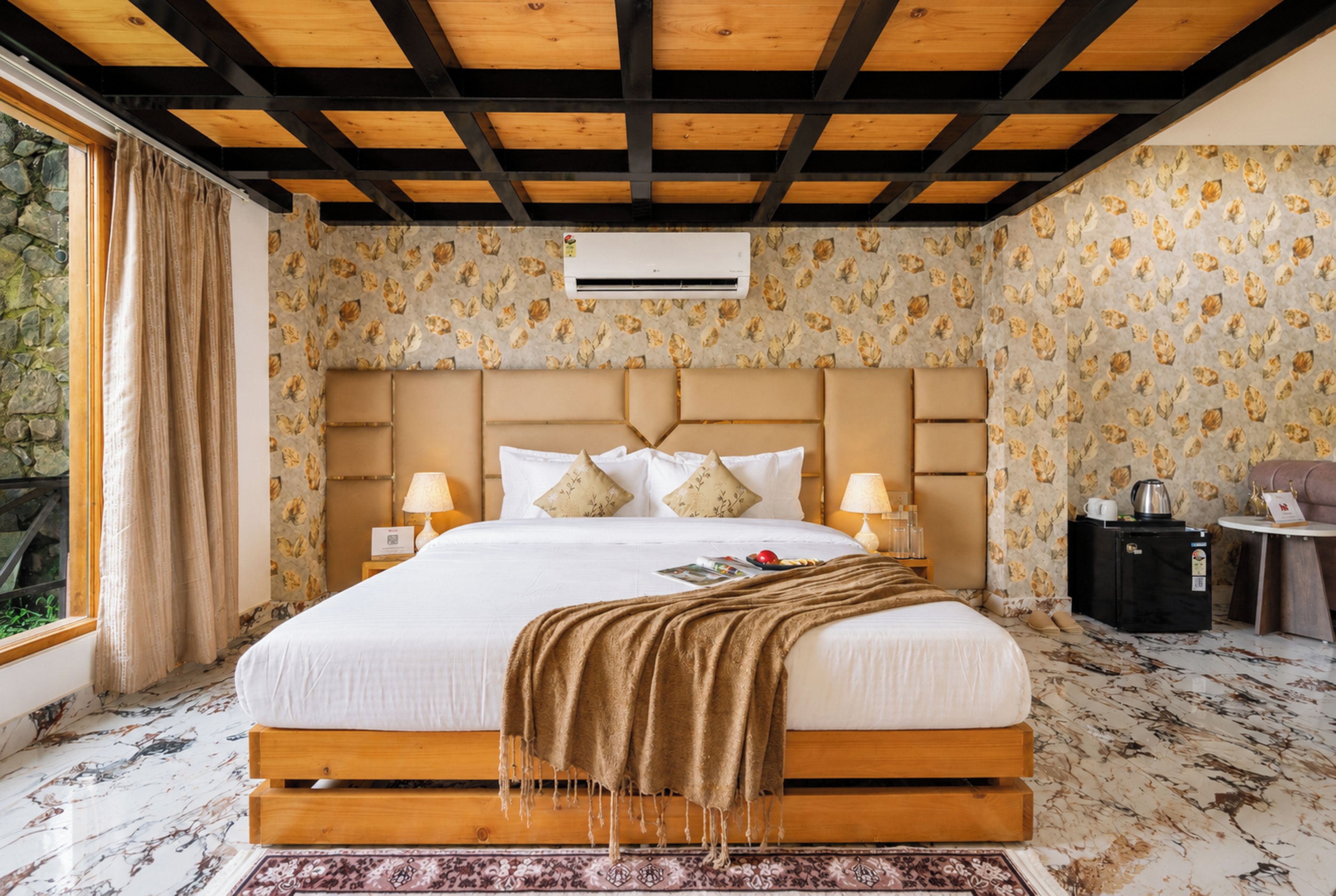 Habitación de hotel con una cama grande, cabecera, dos mesitas de noche con lámparas, una pequeña mesa con hervidor y una ventana con cortinas.