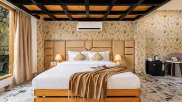 Habitación de hotel con una cama grande, cabecera, dos mesitas de noche con lámparas, una pequeña mesa con hervidor y una ventana con cortinas.