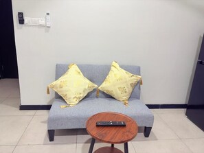 Ein graues Sofa mit zwei gelben Kissen mit Muster, ein kleiner runder Holztisch mit einer Fernbedienung und ein an der Wand befestigter Lichtschalter.