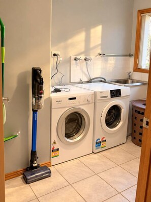 A laundry room with two front-loading washing machines, a vacuum cleaner, and a sink. - Wallcrest Retreat | 5BR, Views, Pets & Parking (Berriedale)