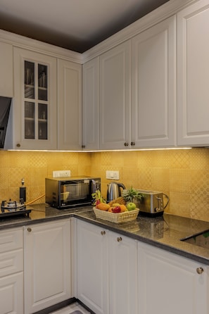 A kitchen with white cabinets, a microwave, a kettle, and a fruit basket on the counter. - ELIVAAS Signature Mistwood 4-BHK (Parra)