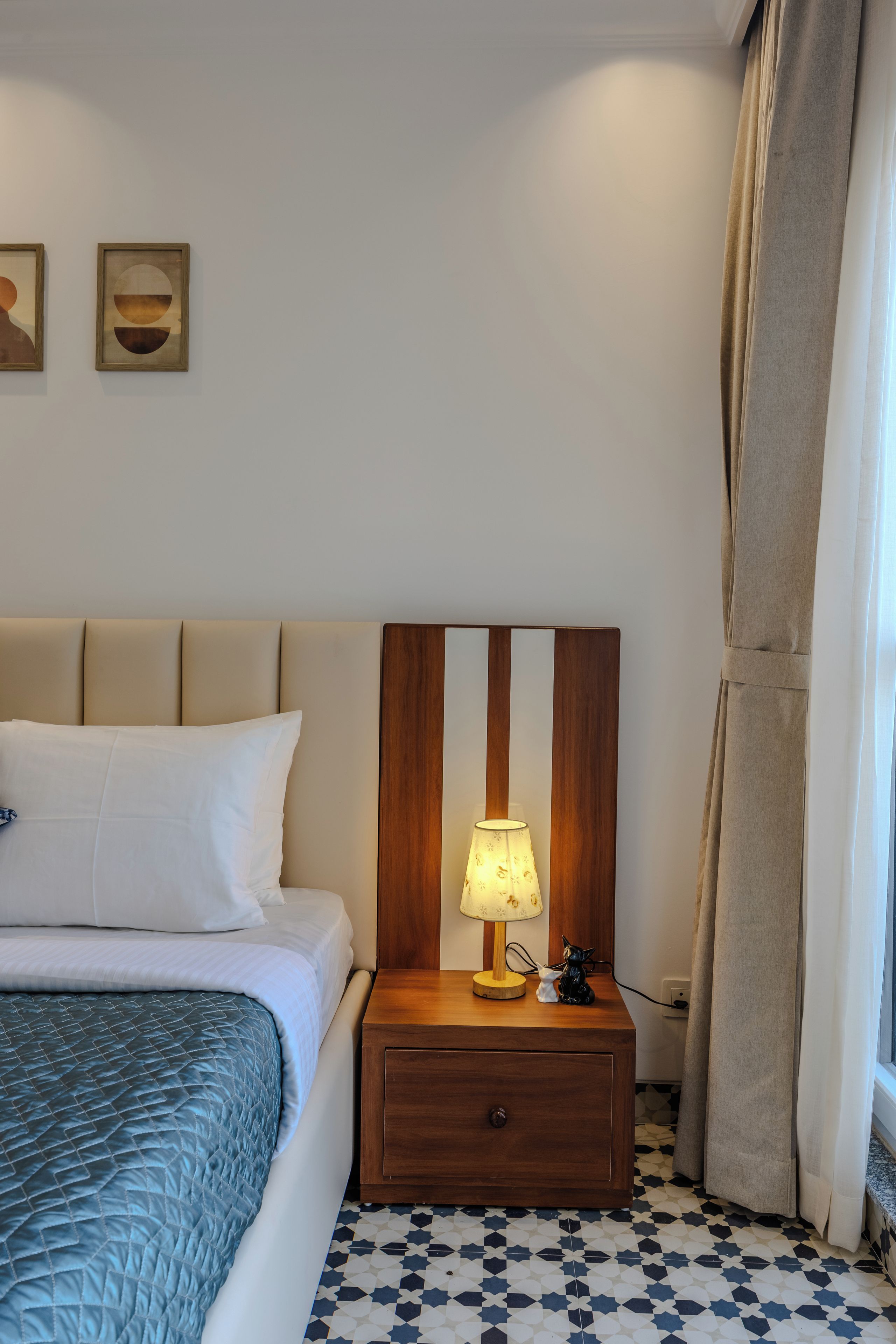 A hotel room with a bed, a nightstand with a lamp, a small statue, and a patterned floor.