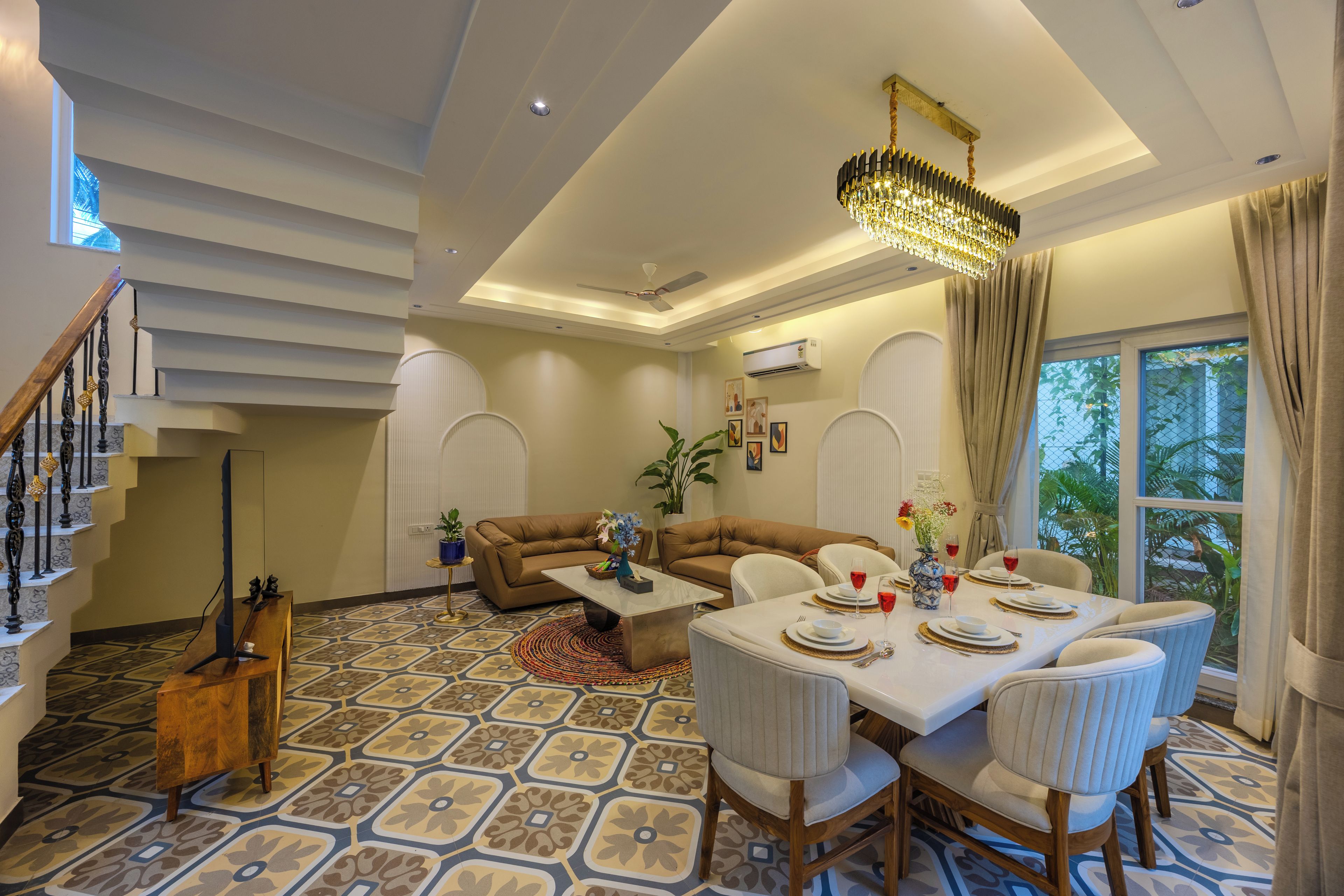 A spacious living area with a high ceiling, a chandelier, a dining table set for four, and a staircase with a decorative railing.