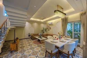 A spacious living area with a high ceiling, a chandelier, a dining table set for four, and a staircase with a decorative railing.