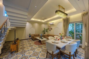 A spacious living area with a high ceiling, a chandelier, a dining table set for four, and a staircase with a decorative railing.