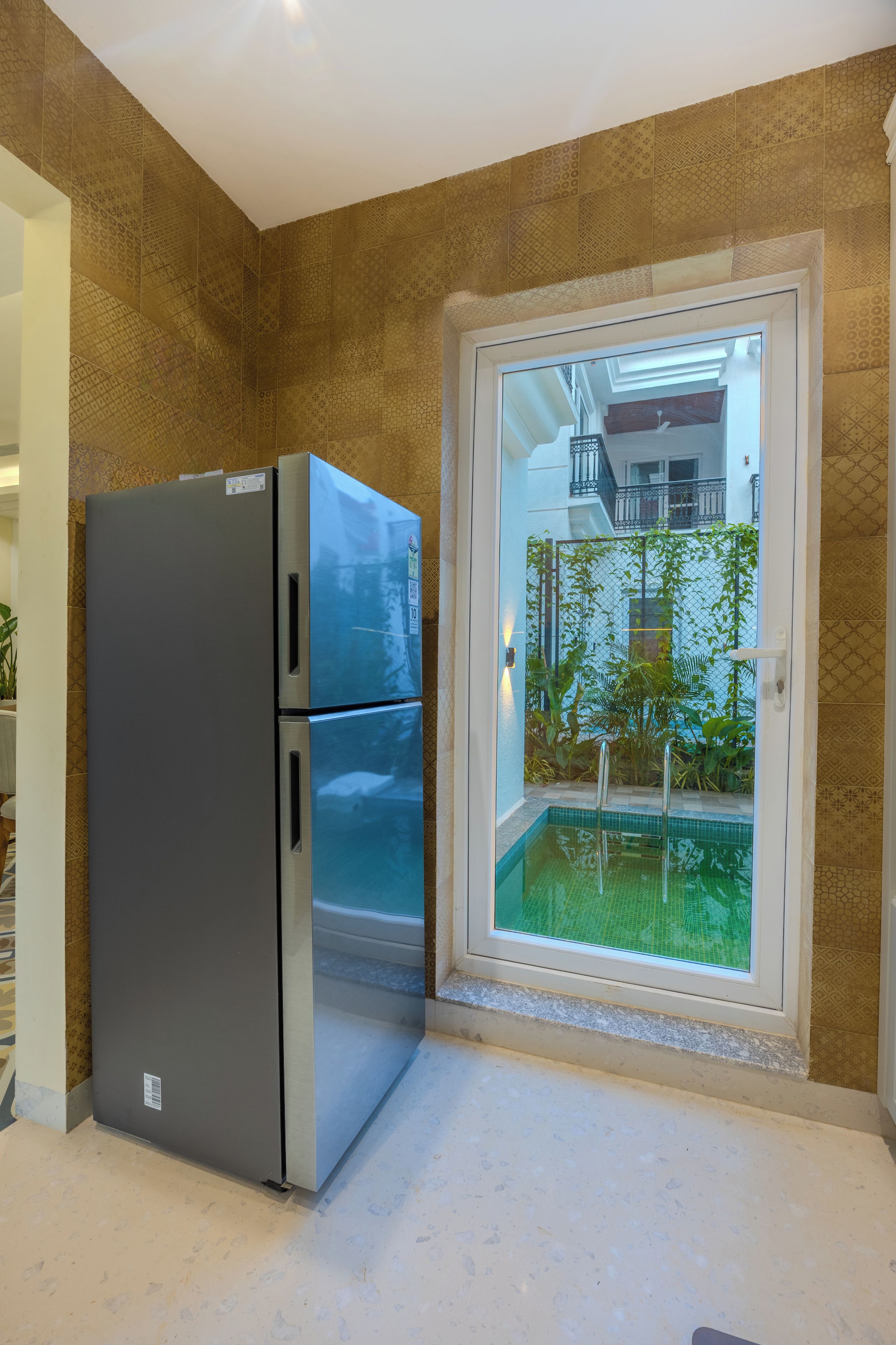 A kitchen with a double-door refrigerator, a window with a view of a pool and greenery, and patterned wall tiles.