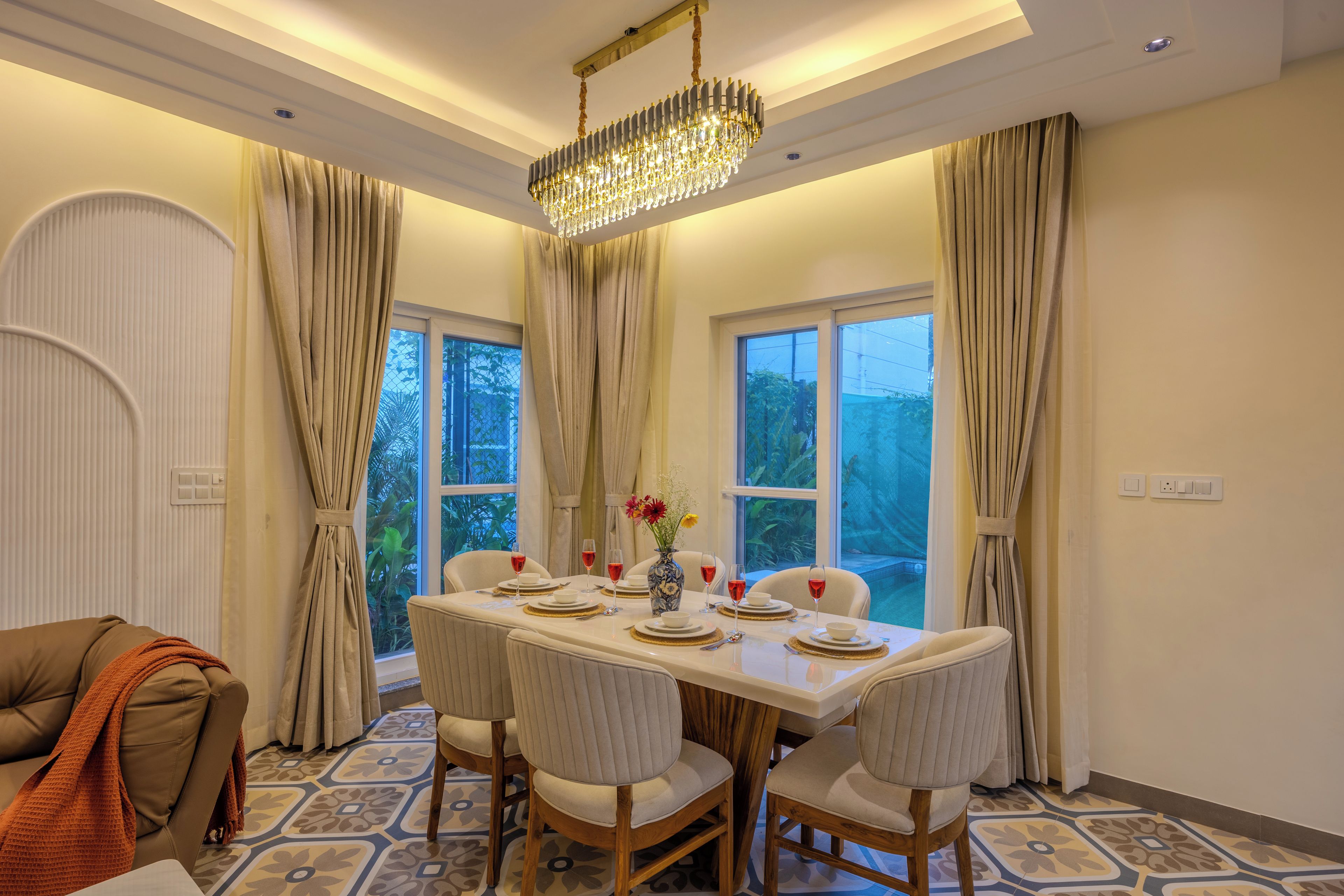 A dining area with a table set for four, surrounded by chairs, with a chandelier above and curtains on the windows.