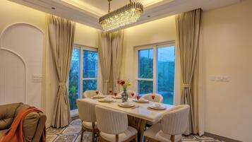 A dining area with a table set for four, surrounded by chairs, with a chandelier above and curtains on the windows.
