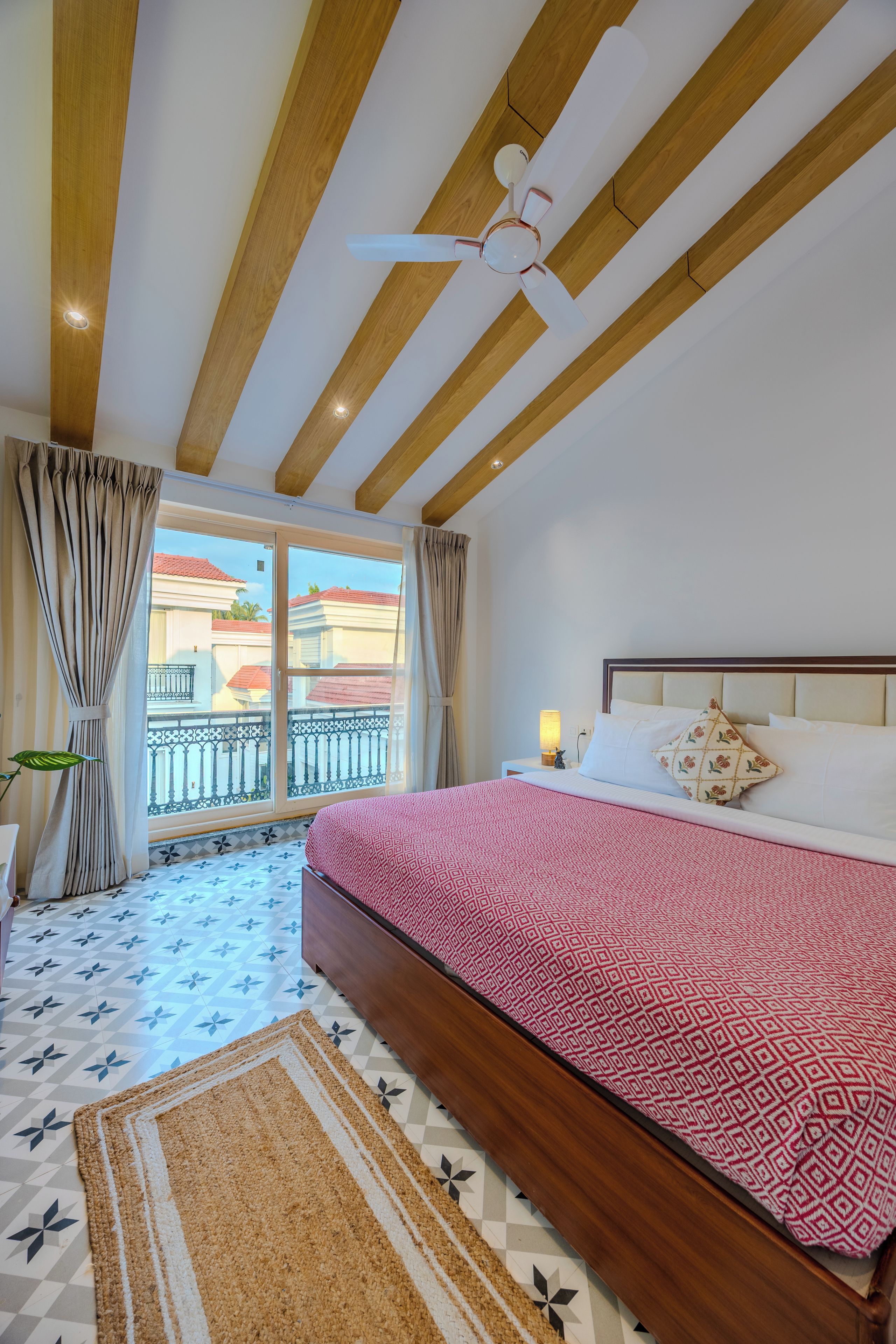 Un dormitorio con cama, ventilador de techo, ventana con cortinas y vista a edificios.