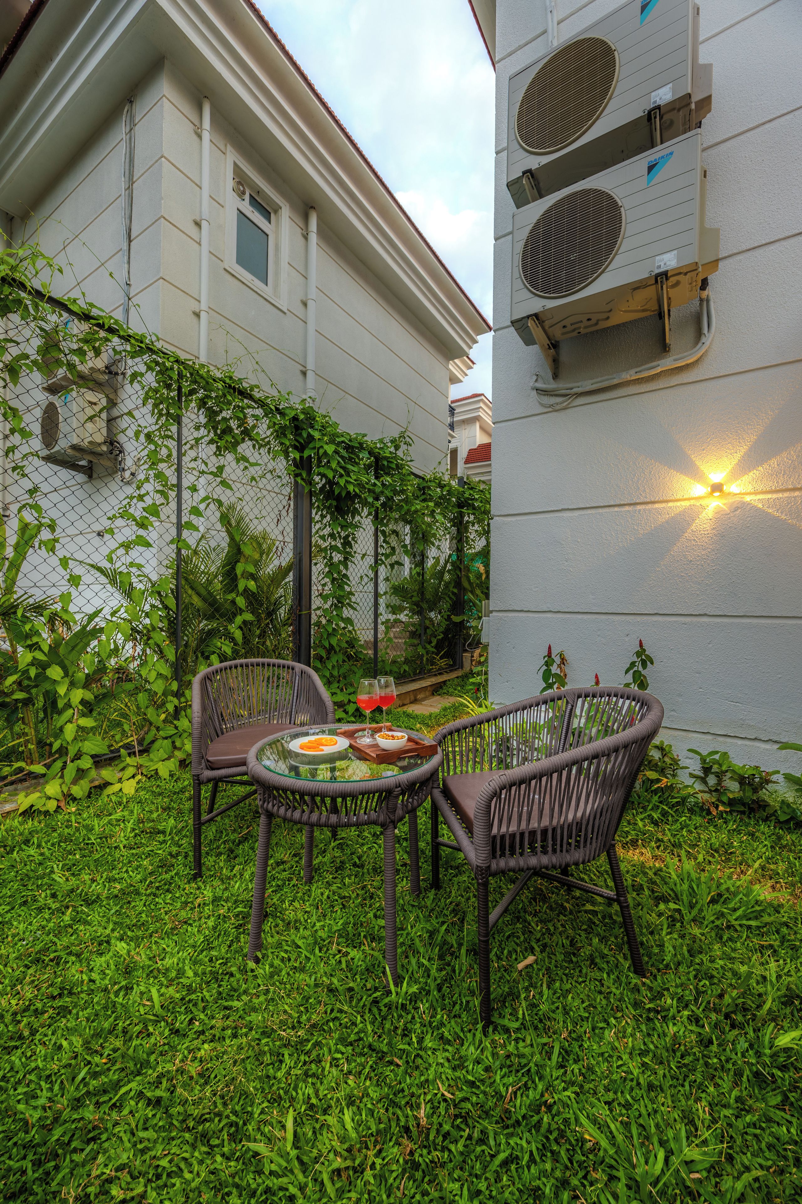 An outdoor seating area with wicker chairs and a glass table, surrounded by lush greenery and a white building with air conditioning units.