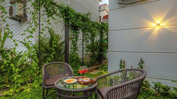 An outdoor seating area with wicker chairs and a glass table, surrounded by lush greenery and a white building with air conditioning units.