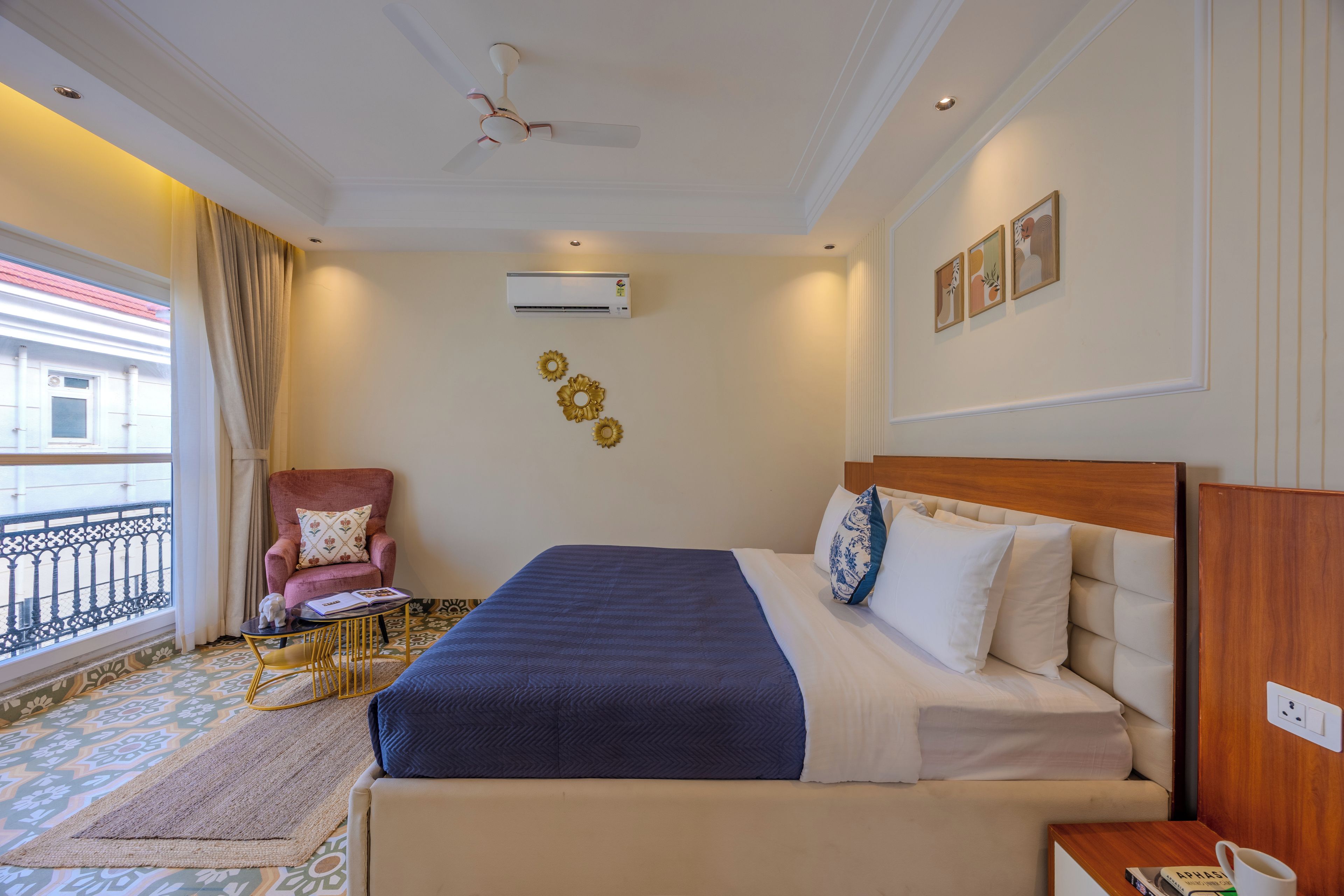 A hotel room with a bed, a chair, a small table, an air conditioning unit, and a window with a view of a building.