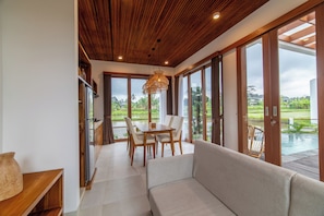 A modern interior with a dining area, sofa, and large windows offering a view of a garden and pool. - SagaLuna Private Villa (gianyar)