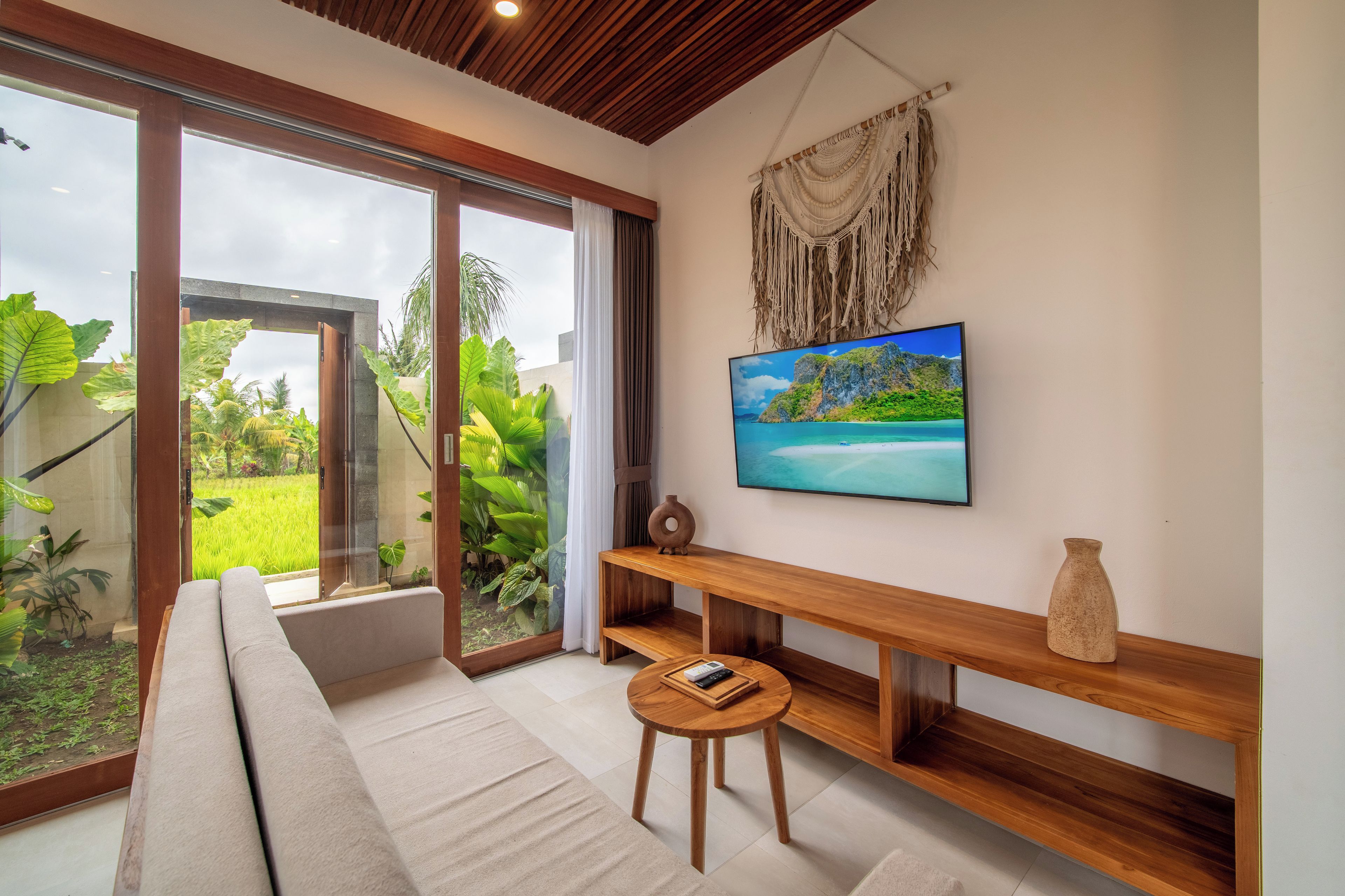 A modern living room with a large sliding glass door leading to a lush garden, a wooden bench, a small round table, and a flat-screen TV mounted on the wall.