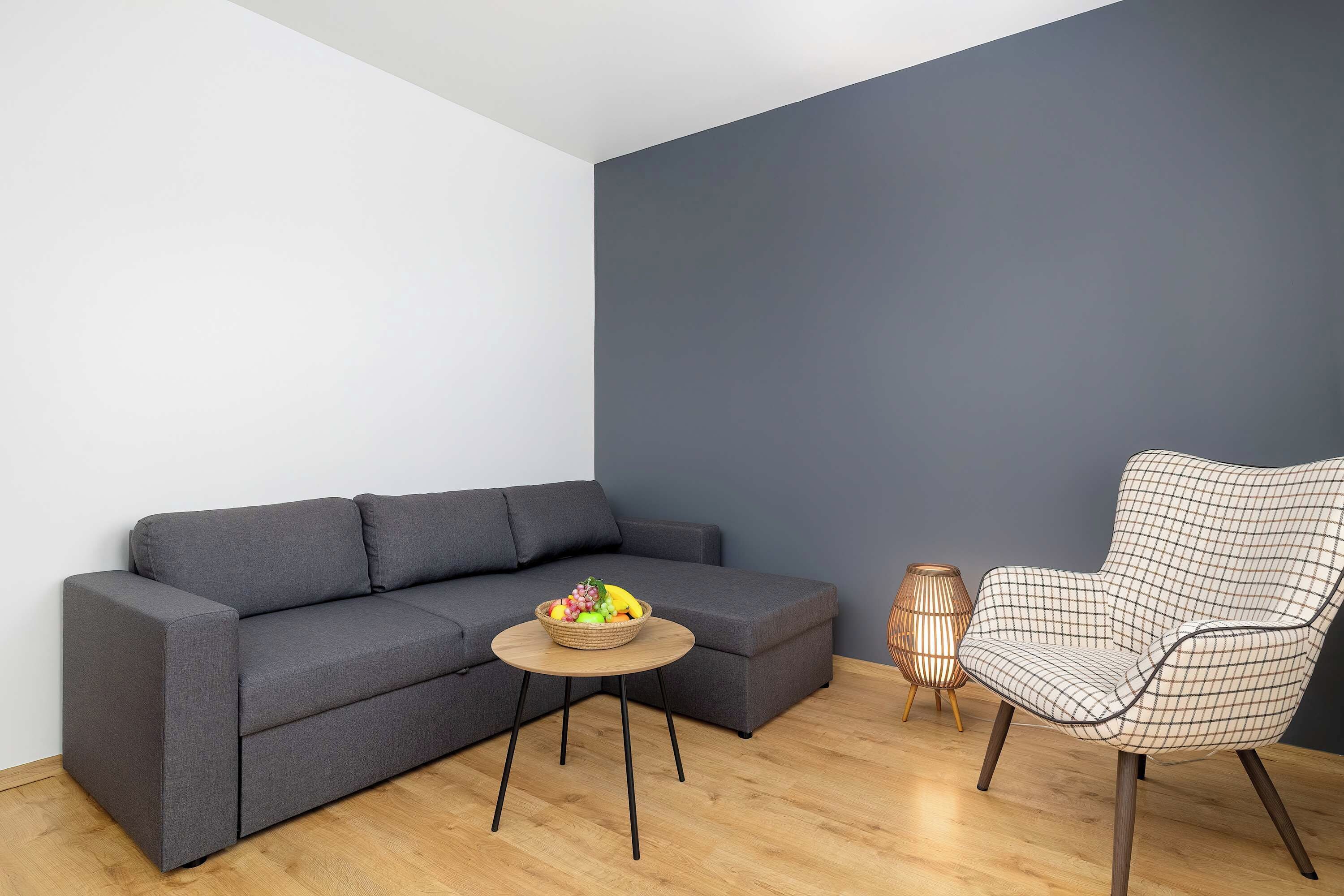 A modern living room with a grey sofa, a round wooden coffee table with fruit, a checkered armchair, and a dark blue accent wall.