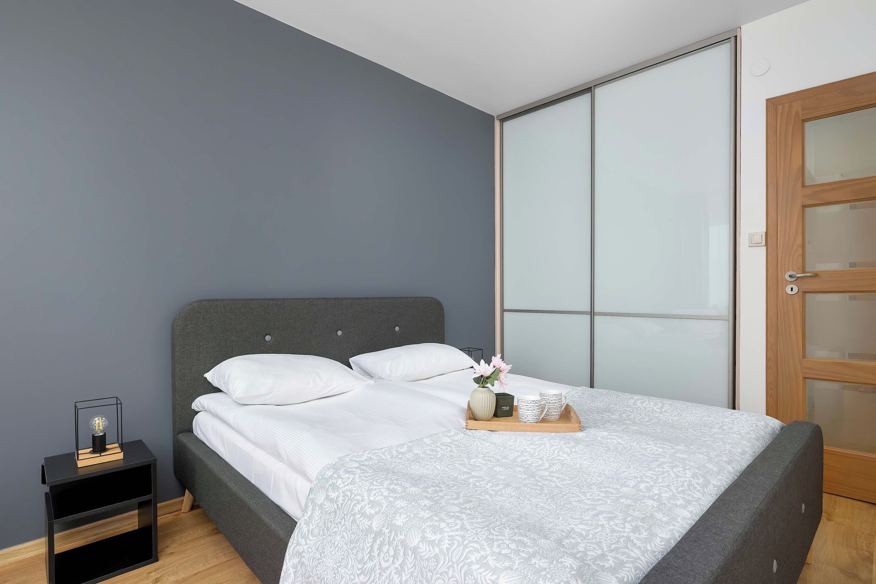 Une chambre moderne avec une tête de lit grise, du linge de lit blanc, une table de chevet avec une lampe et une porte en bois dotée de panneaux en verre dépoli.
