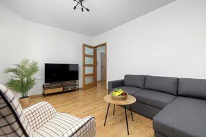Una sala de estar moderna con un televisor de pantalla plana, un sofá gris, un sillón estampado, una mesita redonda con fruta y una planta en maceta.
