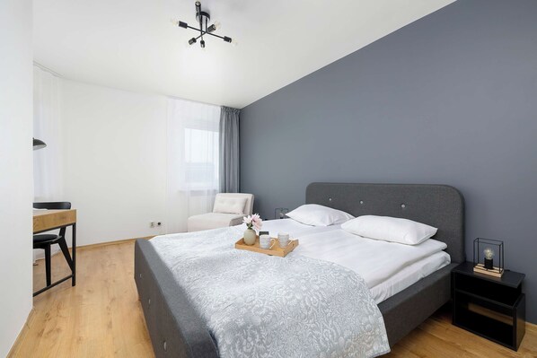 Un dormitorio moderno con una cama tapizada en gris, un escritorio de madera, una silla y una mesita de noche con lámpara.