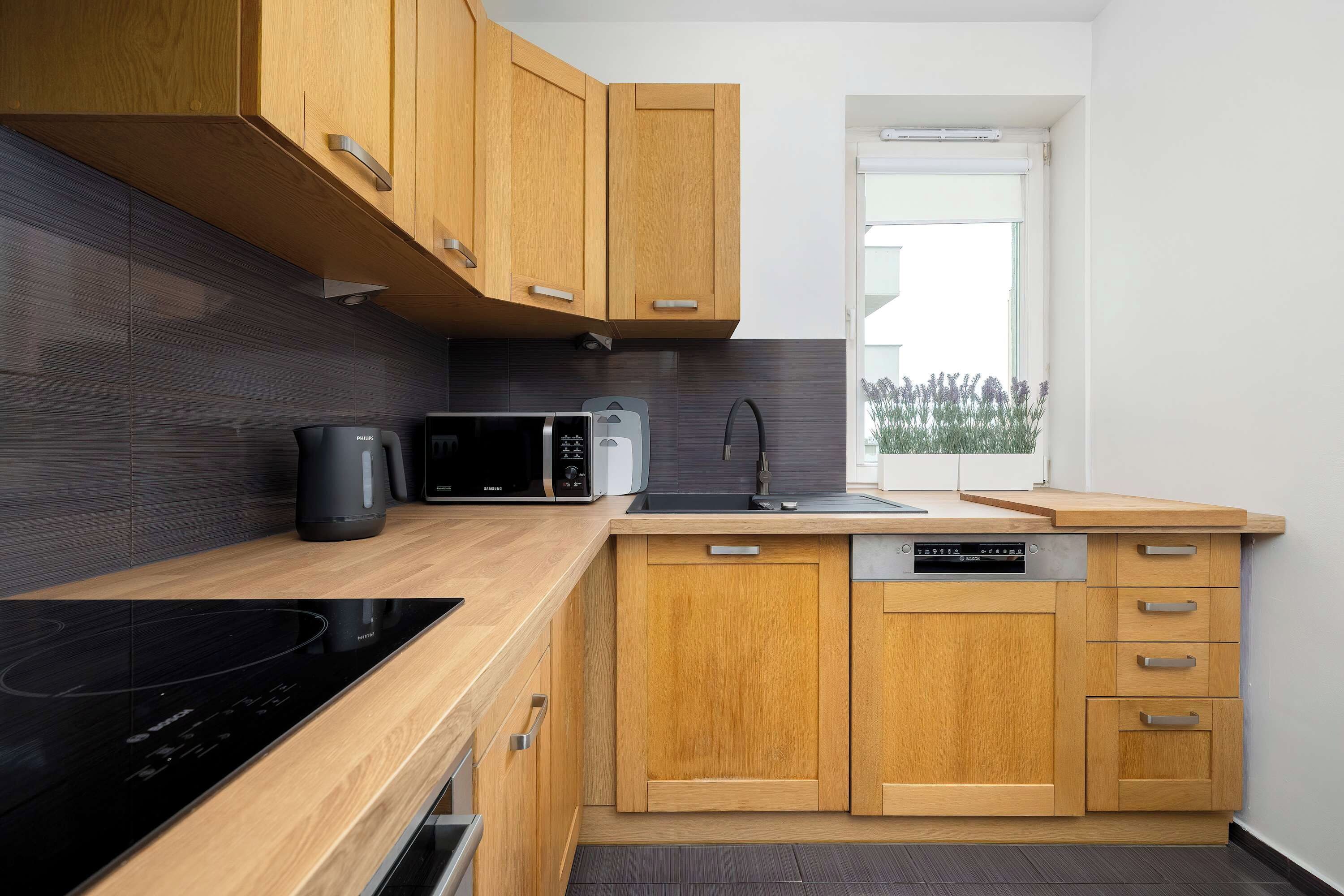 Une cuisine moderne avec des meubles en bois, une plaque de cuisson à induction noire, un four à micro-ondes, un lave-vaisselle et une fenêtre donnant sur l’extérieur.