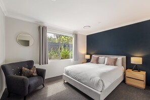 Ein Schlafzimmer mit einem Bett, einem Sessel, einem Nachttisch mit Lampe, einem Spiegel und einem Fenster mit Vorhängen.