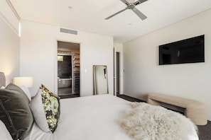 A hotel room with a bed, a ceiling fan, a TV mounted on the wall, a closet with a mirror, and a fluffy white ottoman. - Bayview on Smith—Poolside Sanctuary & Ocean Vistas (Dunsborough)