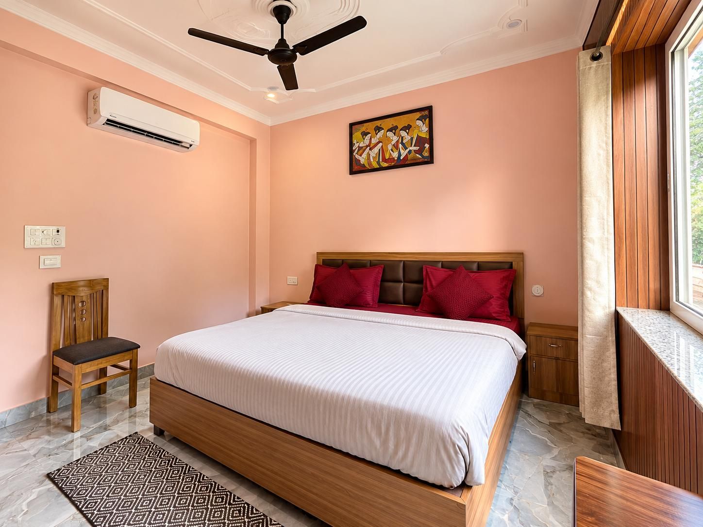 A bedroom with a bed, a ceiling fan, an air conditioning unit, a painting, a window with curtains, and a wooden chair.