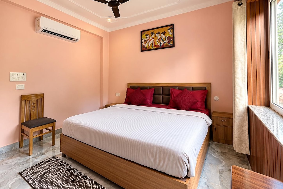 A bedroom with a bed, a ceiling fan, an air conditioning unit, a painting, a window with curtains, and a wooden chair.