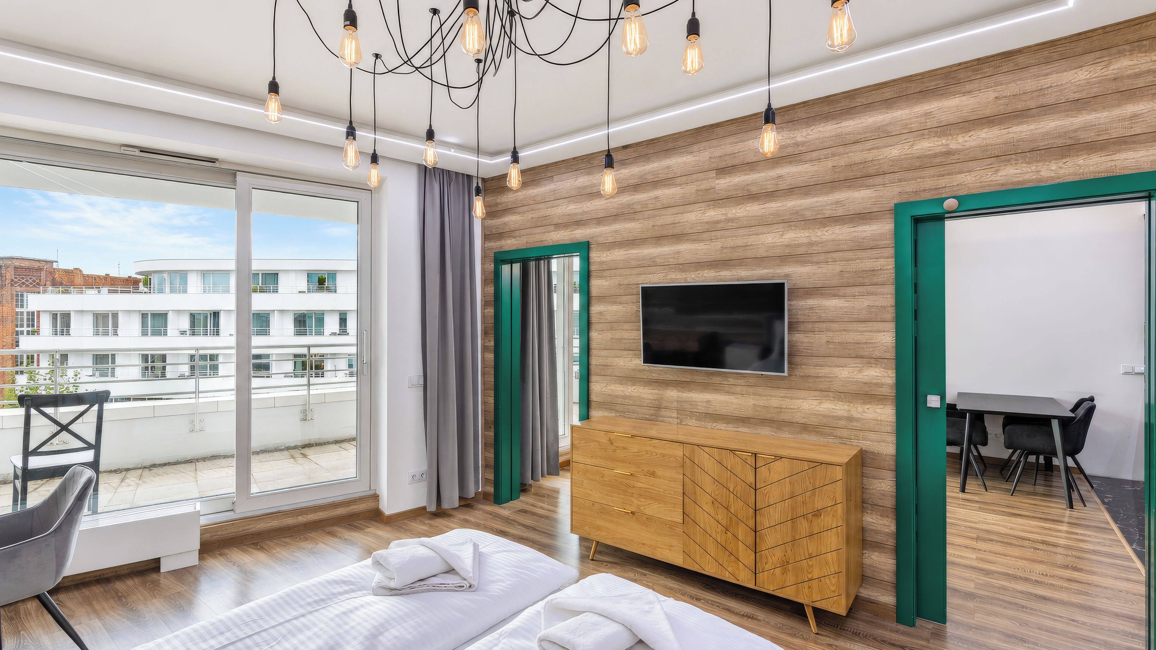A modern hotel room with a large bed, a wooden dresser, a TV mounted on the wall, and a balcony with a view of the city.