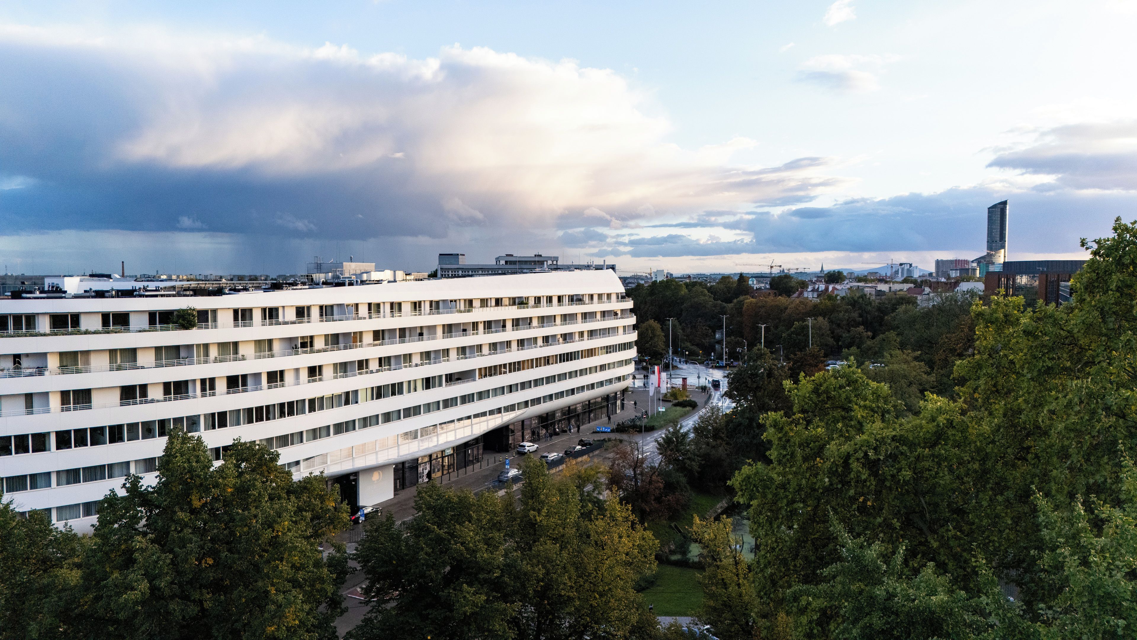 A multi-story building with a flat roof, surrounded by trees and a body of water in the background.