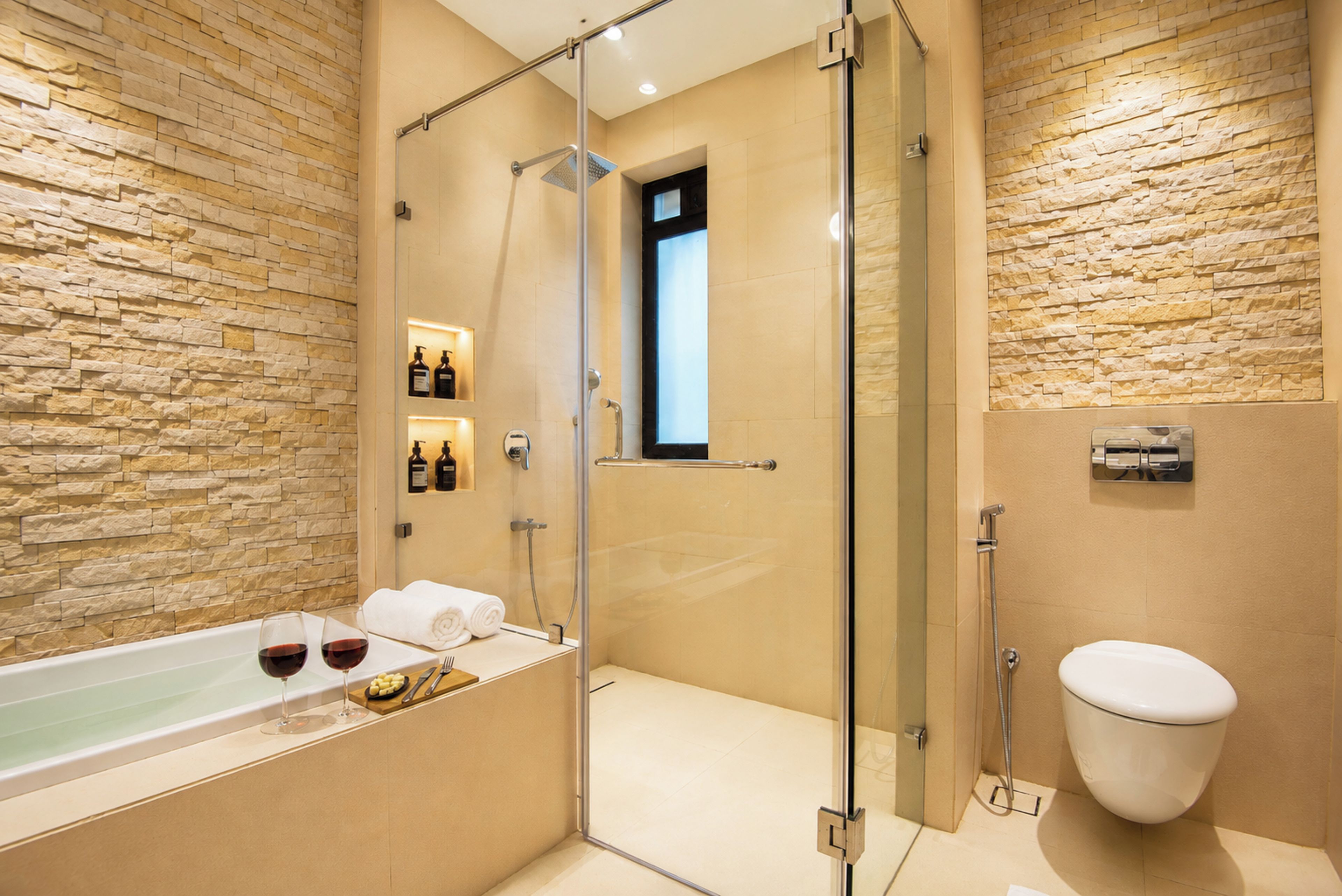 A modern bathroom with a glass-enclosed shower, a bathtub with wine glasses and towels, and a toilet.