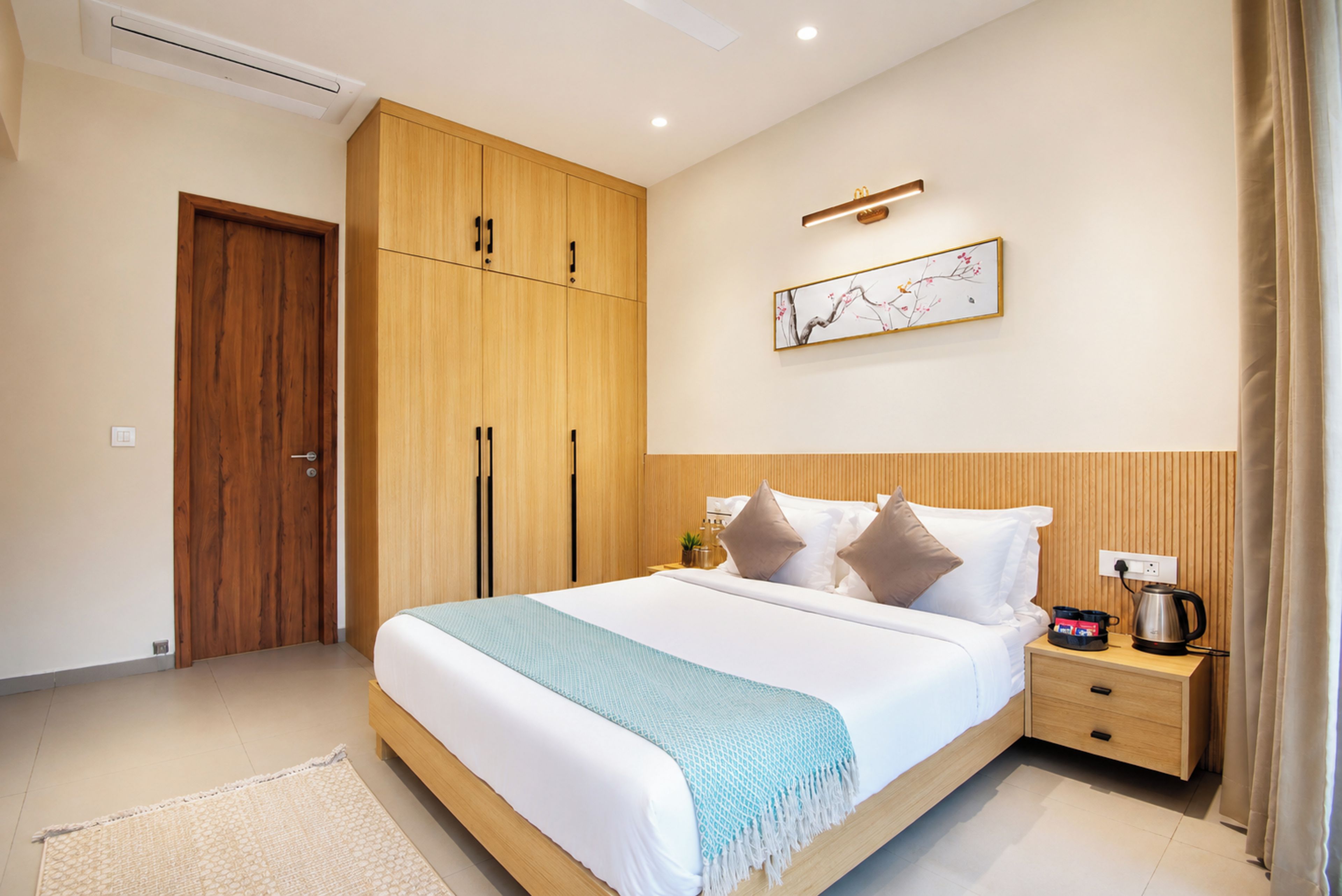 A hotel room with a large bed, a wooden wardrobe, a bedside table with a kettle, and a wall-mounted artwork.