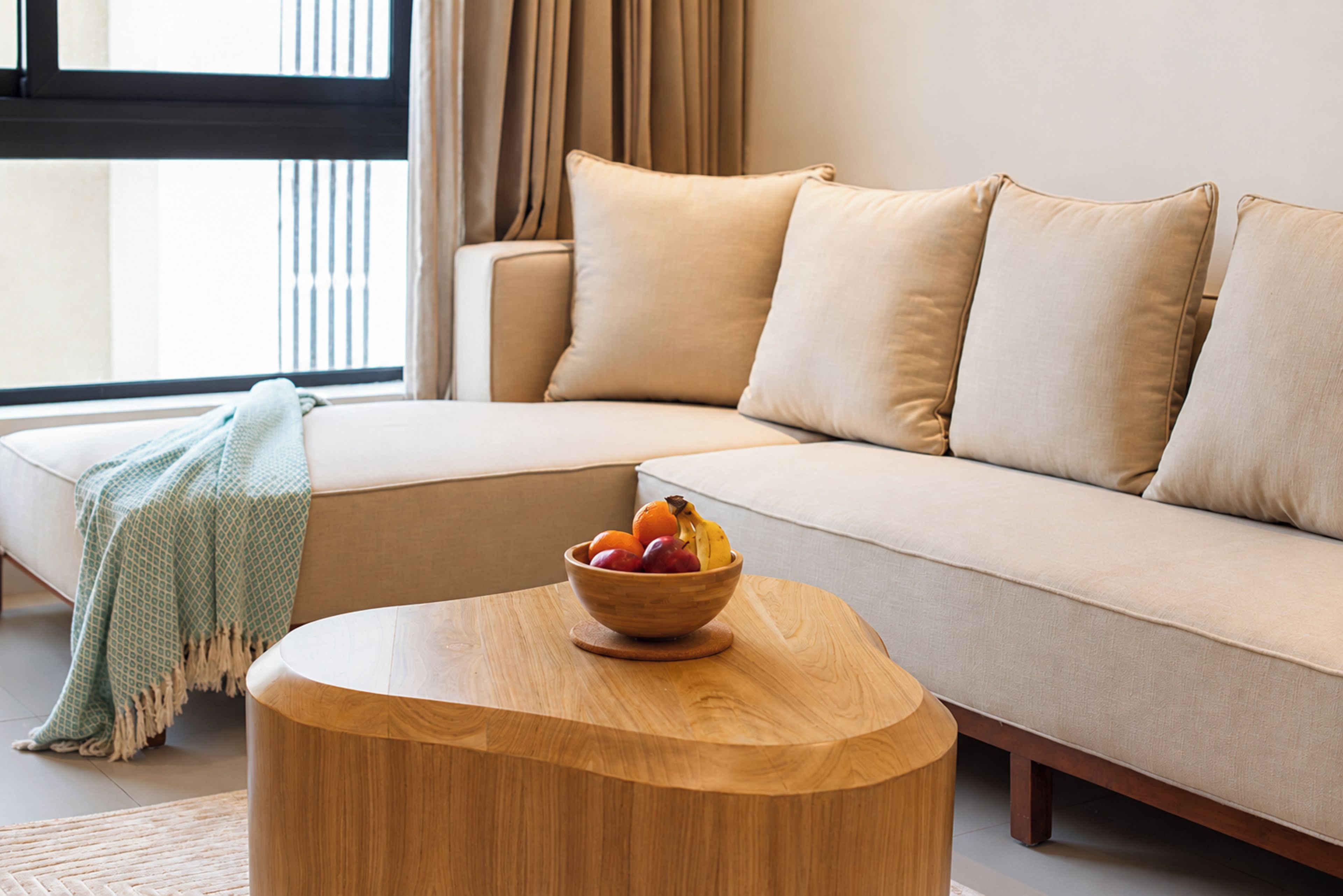 A modern living room with a beige sofa, a round wooden coffee table with a bowl of fruit, and a patterned throw blanket.
