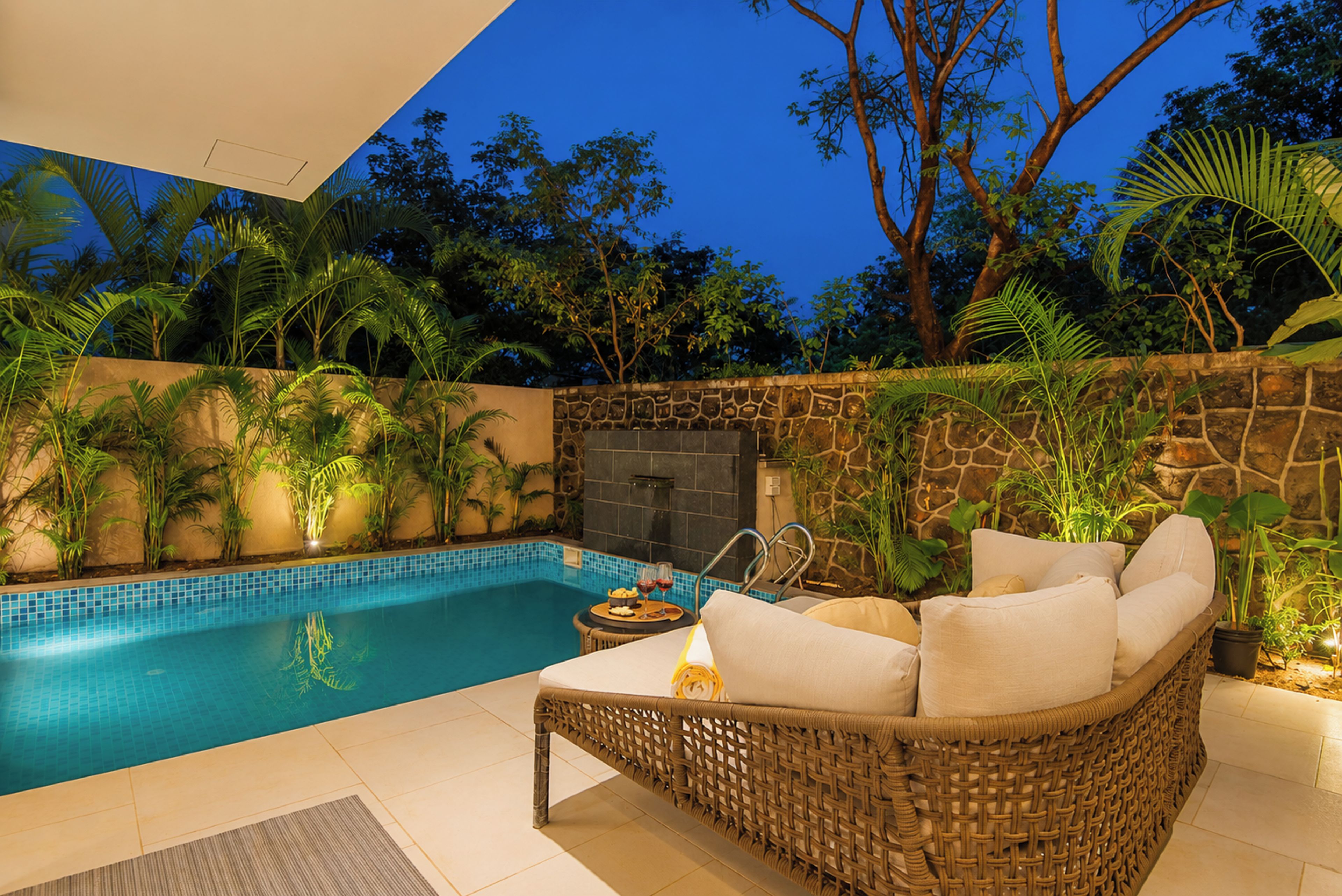 A poolside area with a wicker sofa, a stone wall, and tropical plants.