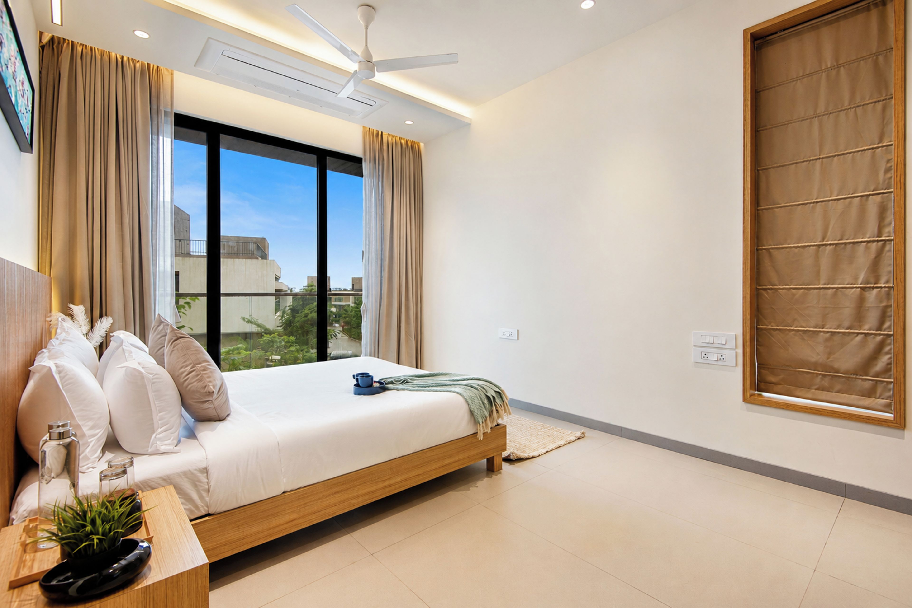Een moderne slaapkamer met een bed, gordijnen, een plafondventilator en uitzicht op gebouwen.