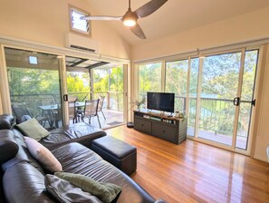 A spacious living area with a leather sofa, a flat-screen TV on a wooden cabinet, and a balcony with a table and chairs. - Heather Beach House - Three Sisters (Point Lookout)