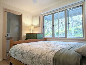 A bedroom with a bed, a nightstand, a lamp, a window with a view of trees, and a door. - Heather Beach House - Three Sisters (Point Lookout)