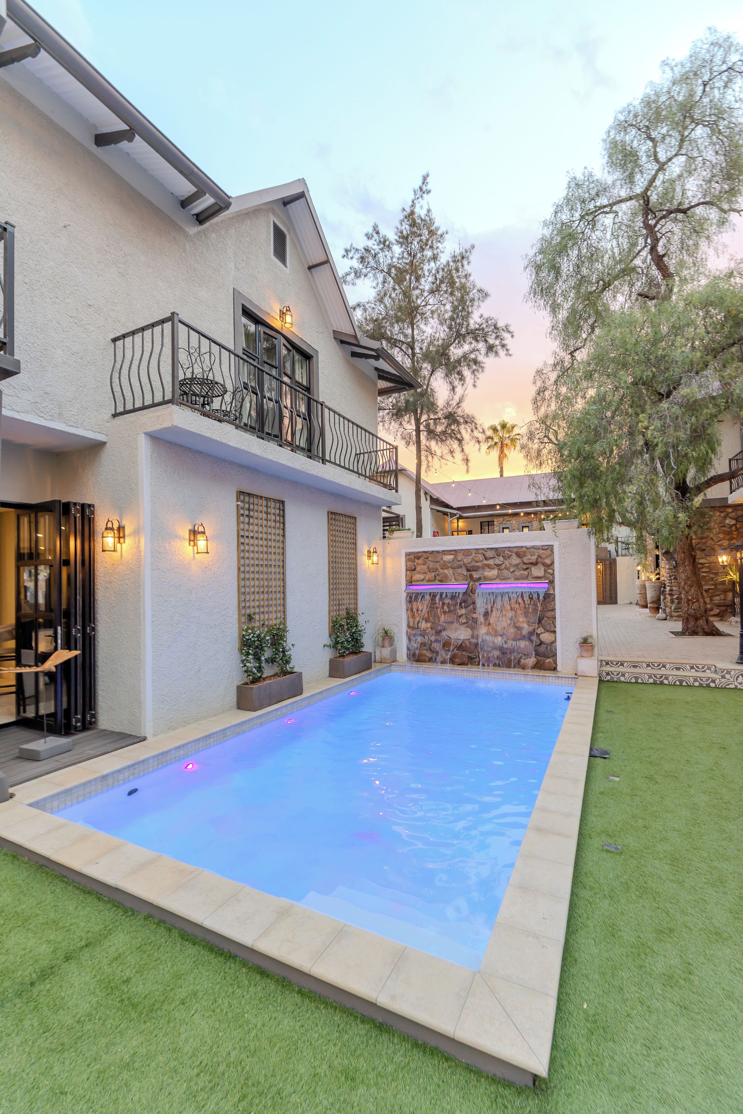 Ein Wohnhaus mit Schwimmbad, einer Steinmauer mit Wasserspiel und einem Balkon mit Gartenmöbeln.
