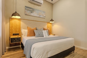 A bedroom with a bed, wooden headboard, bedside tables, and a painting on the wall. - Gracious Whisp Boutique Hotels (Windhoek)